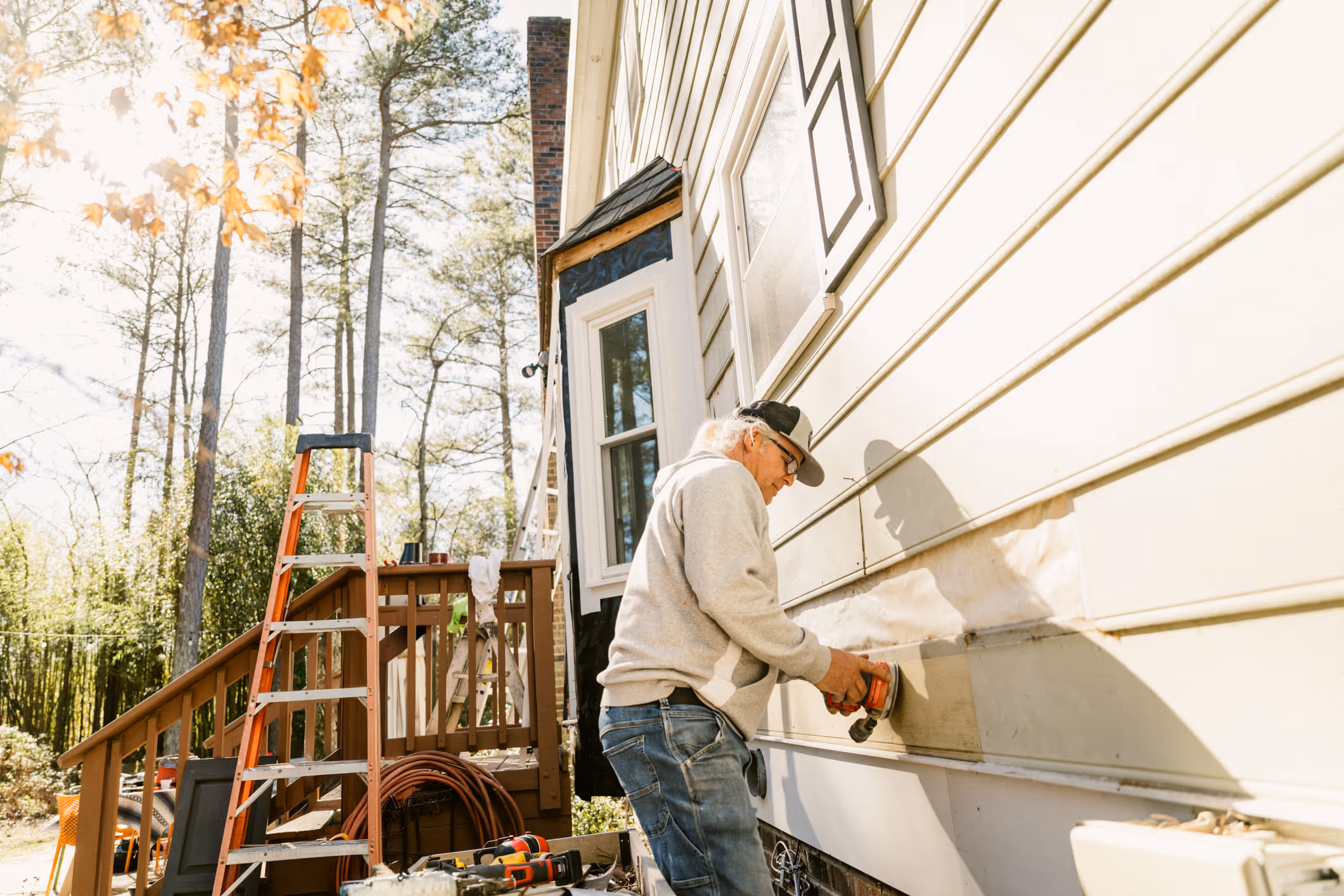 A contractor uses a power tool to repair vinyl siding panels on a residential home exterior, with ladders and equipment visible on the job site.